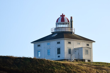 A historic square white and red wooden tower lighthouse with a round light room. There are a number of small windows on all sides of the building.  There's grass on the ground and blue sky in the back