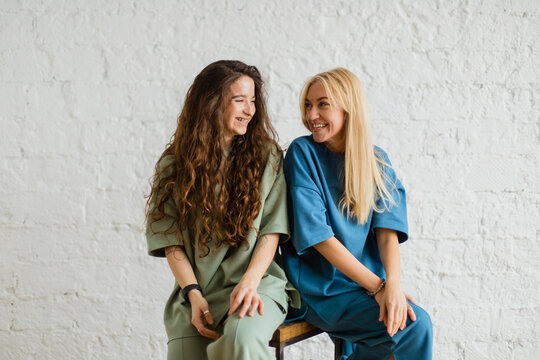 Two girls sit near white wall hugging each other