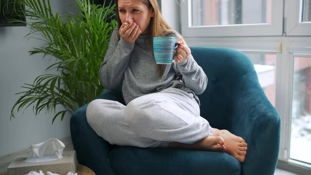 Unhealthy woman sits in a chair, drinks hot tea or a cold medicine and sneezes or blows her nose into a napkin because she has a cold, flu, coronavirus. It is snowing outside