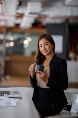 Smiling young Asian businesswoman working on a laptop computer at her desk in a bright modern home office, doing calculating expense financial report finance making notes on paper graph data document.