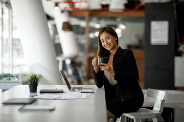 Smiling young Asian businesswoman working on a laptop computer at her desk in a bright modern home office, doing calculating expense financial report finance making notes on paper graph data document.