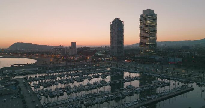 Aerial view of Barcelona skyscrapers in the port and beach area by the sea at sunset. Modern offices, boats and evening lights.
