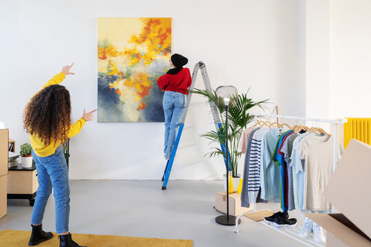 Diverse women hanging painting on wall in house