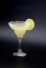 Margarita cocktail, an alcoholic drink in a tall glass with salt around the rim and a slice of lime on a black background.