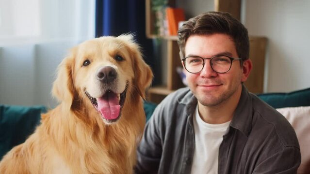 Portrait of young smiling man wearing glasses and his beloved dog sitting on sofa in living room. Golden retriever looking in camera and posing together with male owner. Guy petting his domestic pet.
