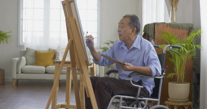 Asian senior man painting picture at living room. He sitting wheelchair while painting picture. Healthcare, Lifestyle and Hobby concept.