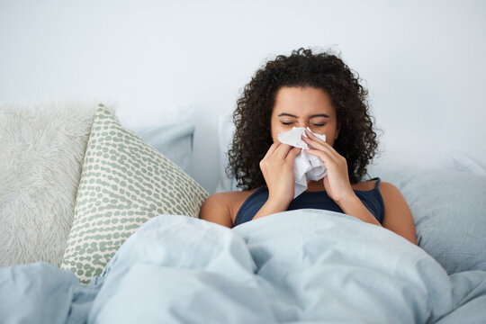 I definitely cant go to work with this cold. Shot of an attractive young woman feeling sick and blowing her nose while in bed in the morning.
