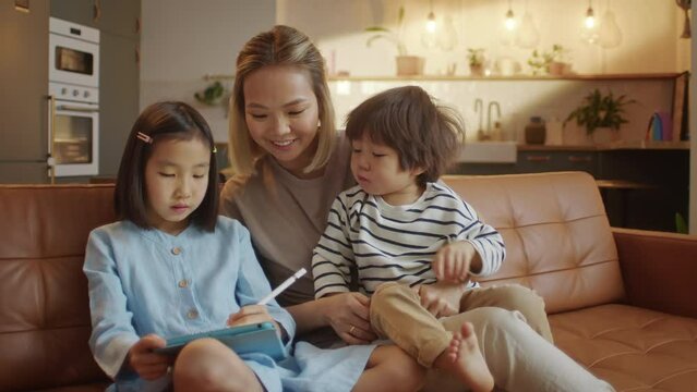 Happy Asian Family Using Tablet Computer With Children Mother Watching Kids Play Game on Touchscreen Technology Sitting on Sofa. Using Education Application for Easy and Funny Learning,