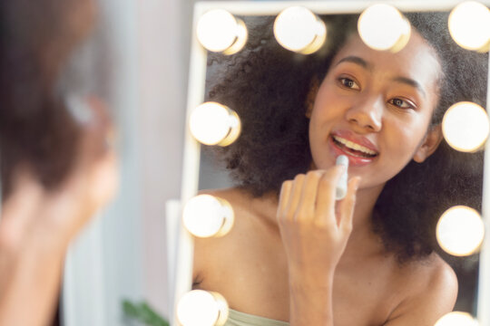 half-Thai-African American girl puts on her own makeup, holds her own lipstick and puts herself in the mirror with side lights in the house