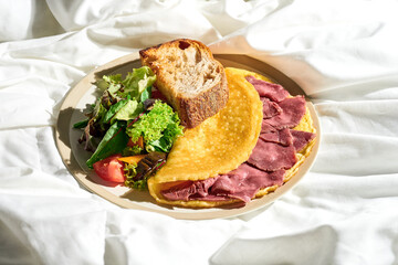 Omelet with roast beef, vegetable salad and toast in a plate on a white sheet. Tough light.