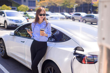 Business  woman standing on city parking near electric car, charging automobile battery from small city station, drinking coffee and using smartphone