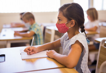 Portrait of diligent schoolgirl wearing protective face mask sitting in class writing exercise, new...