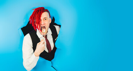 Hipster eats sushi roll. Man with red dreadlocks holding in hand sticks chopsticks with sushi roll traditional japanese food looking at camera through hole in blue wall in studio