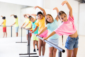 Fototapeta premium Diligent tween girl practicing classic dance moves near ballet barre during group class.