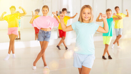 Fototapeta premium Smiling little girl training movements of modern vigorous dance with group of tweens in children dance studio.