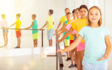 Fototapeta premium Smiling tween girl practicing classic dance moves near ballet barre during group class.