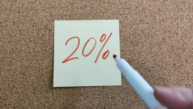 The phrase 20% on a yellow paper sticker. Drawing with a felt-tip pen. A woman's hand with a red marker. Sticker on a cork board close-up