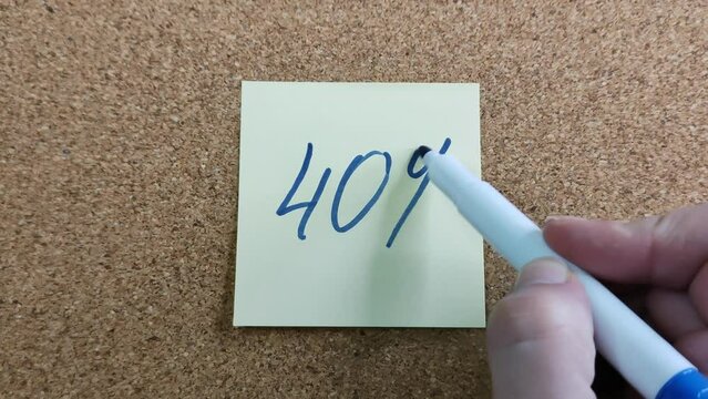 Writing a phrase 40% with a felt-tip pen on a yellow paper sticker. A blue marker in a woman's hand. A sticker on a cork board for notes. Close-up 