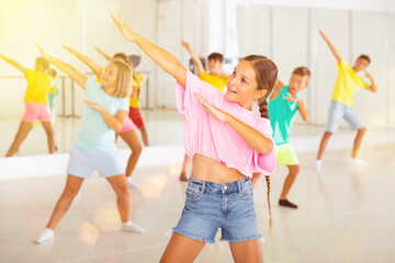 Smiling little girl training movements of modern vigorous dance with group of tweens in children...