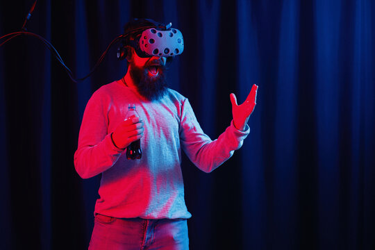 a man with a long black beard in gray golf and virtual reality glasses with a bottle of soda in neon light on a dark background explores the metaverse