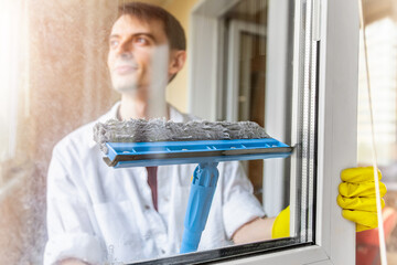 Cleaning concept. Smiling young man washing window in yellow gloves using squeegee or rag, close...