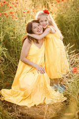 Fototapeta premium A young pregnant woman with her daughter in yellow dresses are standing in a poppy field. Family relationships