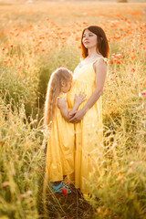 A young pregnant woman with her daughter in yellow dresses are standing in a poppy field. Family relationships