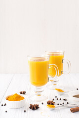 Golden turmeric latte in glasses, spices and cinnamon on white wooden background.