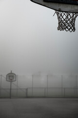 Fototapeta na wymiar Street basketball court with the hoop on focus in the fog conveys sadness and emptiness. Abandoned outdoor basket ball field on a gloomy day provide illustration of war, escape and flight from home