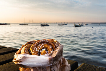 Bag of cinnamon rolls on a wooden boardwalk by the ocean at dusk with the Oresund Bridge in the...