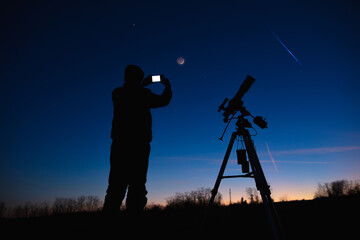 Silhouette of a man with smartphone, telescope and countryside under evening skies. © astrosystem