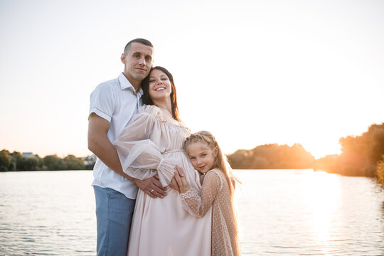 A husband with a pregnant wife and daughter is resting on the river bank, standing on the pier in summer sunny weather at sunset. couple in love man and woman hugging holding hands. A happy family