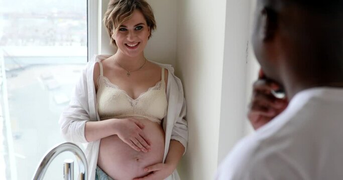 Happy caucasian pregnant woman stands and touches her belly next to african man.