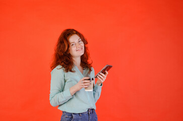 Business portrait of beautiful young woman with red curly hair and blue eyes against red background. Drinking coffee. Using mobile phone. Lifestyle. 