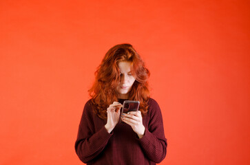 Business portrait of beautiful young woman with red curly hair and blue eyes against red background. Drinking coffee. Using mobile phone. Lifestyle. 