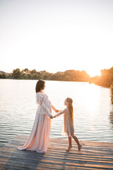 Fototapeta na wymiar A pregnant woman with her daughter against the background of water at sunset, a woman with a belly enjoys the sunset by the river