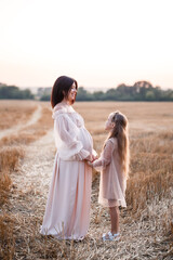 Young pregnant woman with her daughter, happy family hugging on a golden field in the countryside. Family relationships.