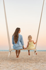 Young mother and little fair-haired girl sit on swing on sea background during sunset. Mom and daughter on the beach over yellow magic sunset lights.