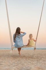 Mom and little girl sit on swing and watch the stunning sunset over the sea. Magical sunrise over sea.