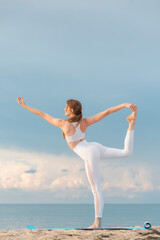Fototapeta premium Young fit woman in white sportswear practices yoga. Natarajasana pose on the beach on bright sunny day.