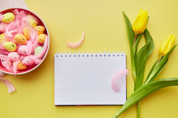 Naklejka na ściany i meble Festive notepad mock up. Pink bowl with colorful eggs, pink feathers and yellow tulips. Happy Easter concept. Post card mock up on yellow background.