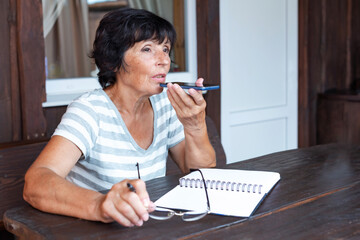 Elderly woman sit at homeoffice room sends a voice message using the phone to client, chatting with...