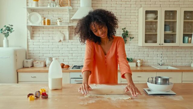 Pretty african american woman with curly fluffy hair rolling out dough with a rolling pin for baking sweet cookies at home in her kitchen.