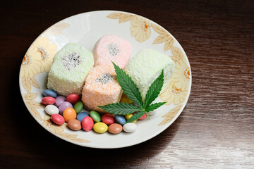 Cannabis dessert. Turkish delight with coconut flakes and colorful candy confetti, top view. Green cmarijuana leaves on white plate background. Layout of hemp food with CBD oil.