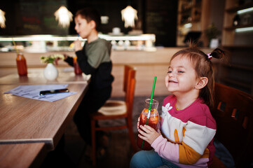 Funny baby girl sitting in cafe and drink juice lemonade.