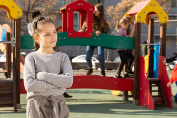 Portrait of upset little girl standing in front of children playing together on playground