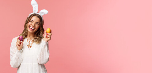 Holidays, spring and party concept. Cheerful good-looking blond woman celebrating Easter day in rabbit ears, holding two painted eggs and wink camera, smiling happily, pink background