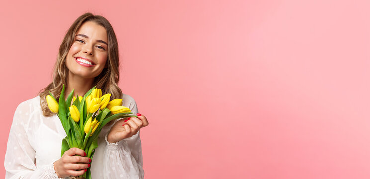 Spring, happiness and celebration concept. Close-up of lovely happy young blond girl in white dress, daydreaming after perfect date, holding yellow tulips and smiling at camera, pink background