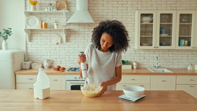 Cute african american woman with curly fluffy hair is whisking eggs with a blender in a bowl while preparing dough for sweet baking cookies at home in her kitchen.