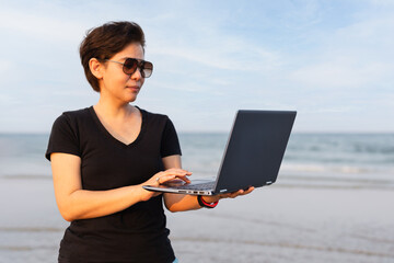 Woman shot hair standing on the beach near the ocean holding a laptop in her hand.